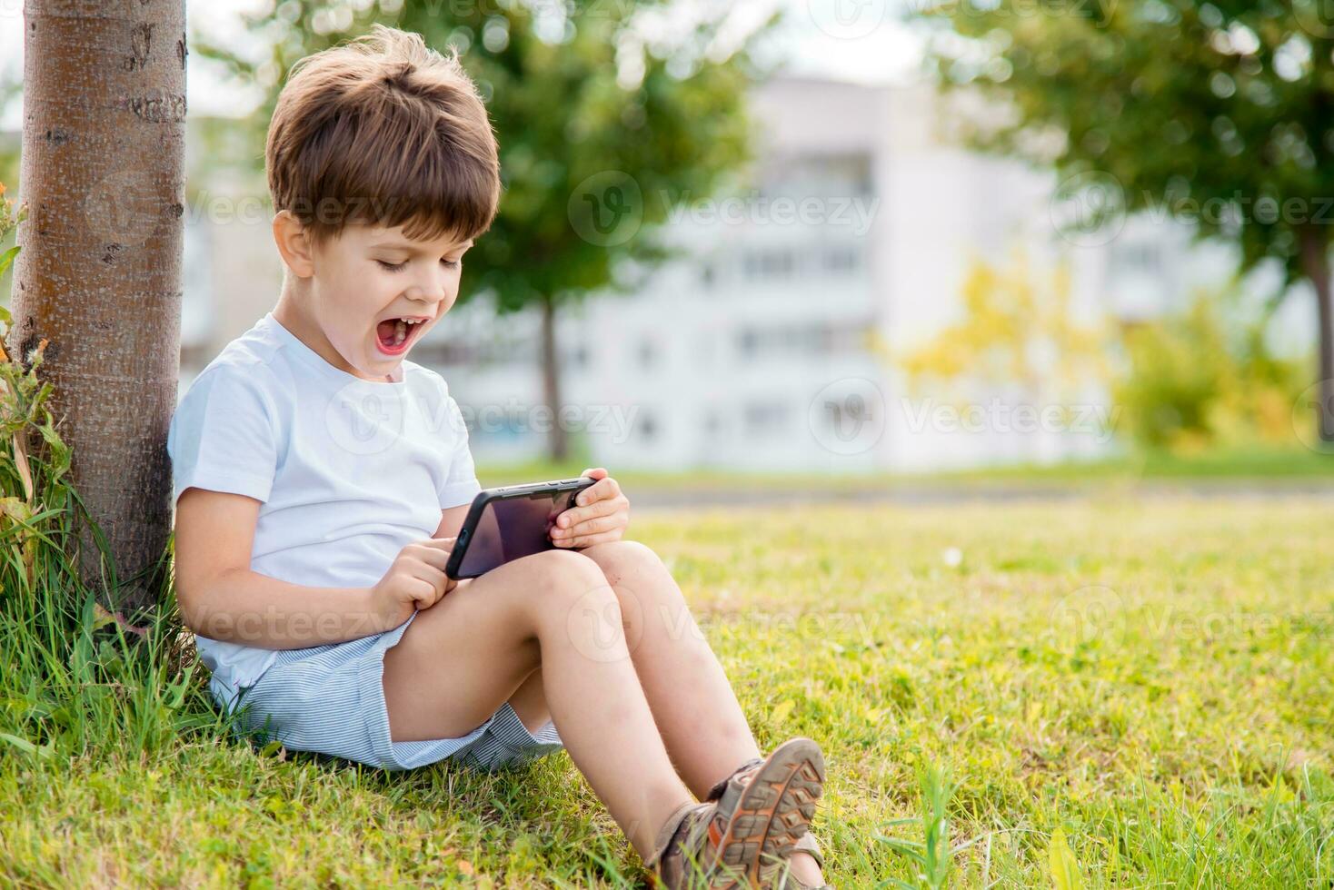 allegro bambino seduta su il erba sembra cartoni animati nel il Telefono nel il estate a tramonto. carino ragazzo avendo divertimento nel natura foto