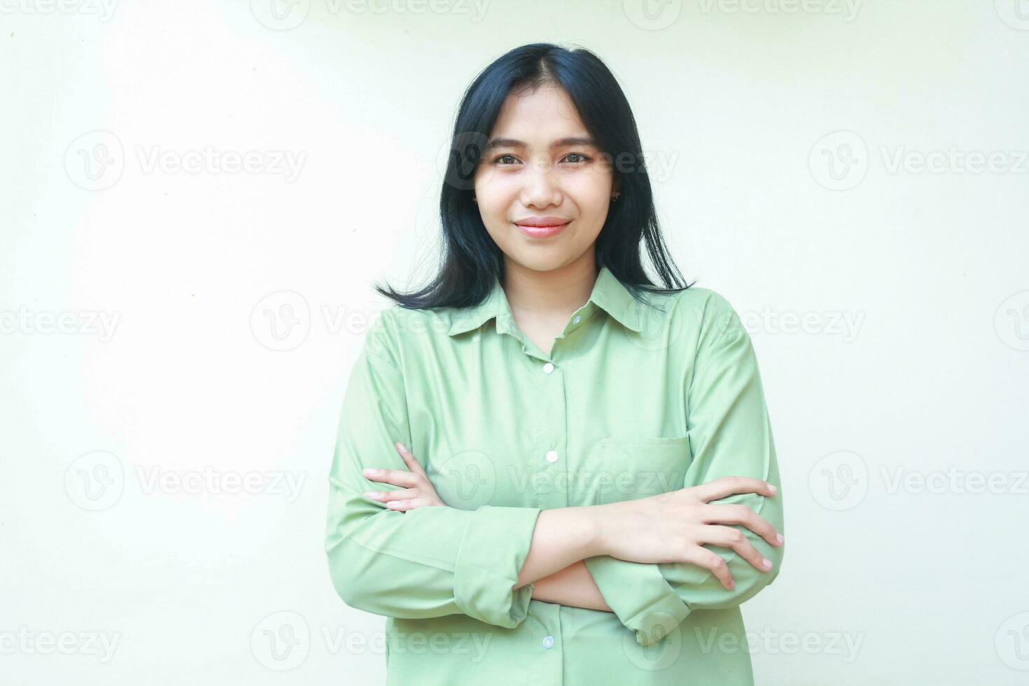 lieta riuscito buio capelli asiatico donna indossare verde al di sopra di dimensione camicia attraversare mani e guardare a telecamera con fiducioso viso espressione in piedi al di sopra di isolato bianca sfondo foto