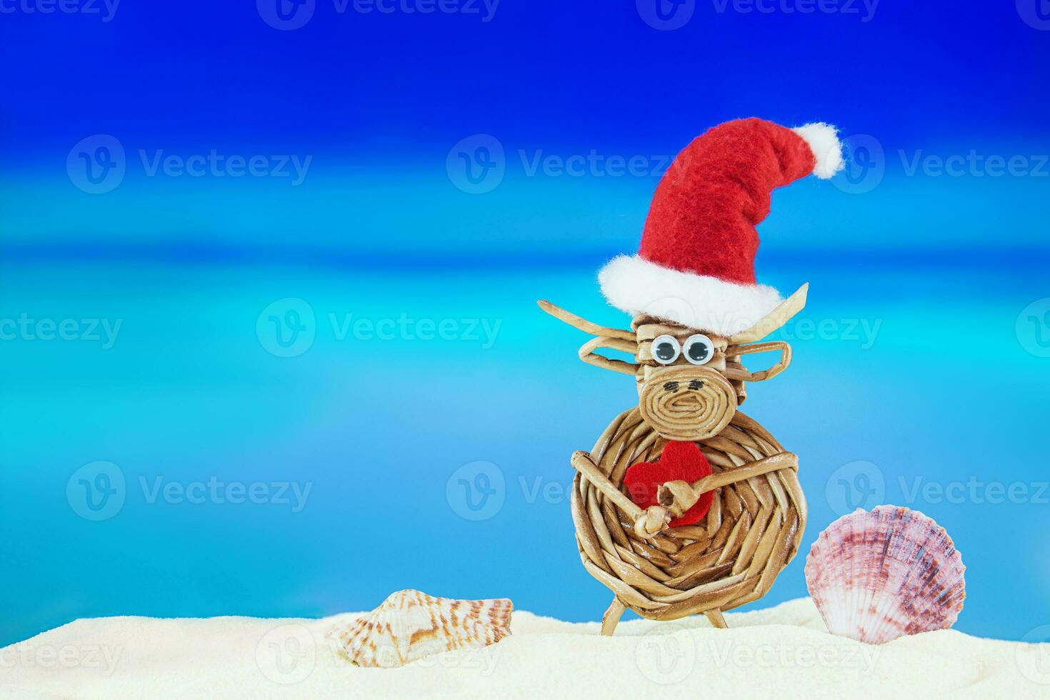 giocattolo intrecciata mucca con babbo natale cappello con cuore e conchiglie su sabbioso spiaggia con mare. Natale, nuovo anno, San Valentino giorno. caldo Paesi. copia spazio foto