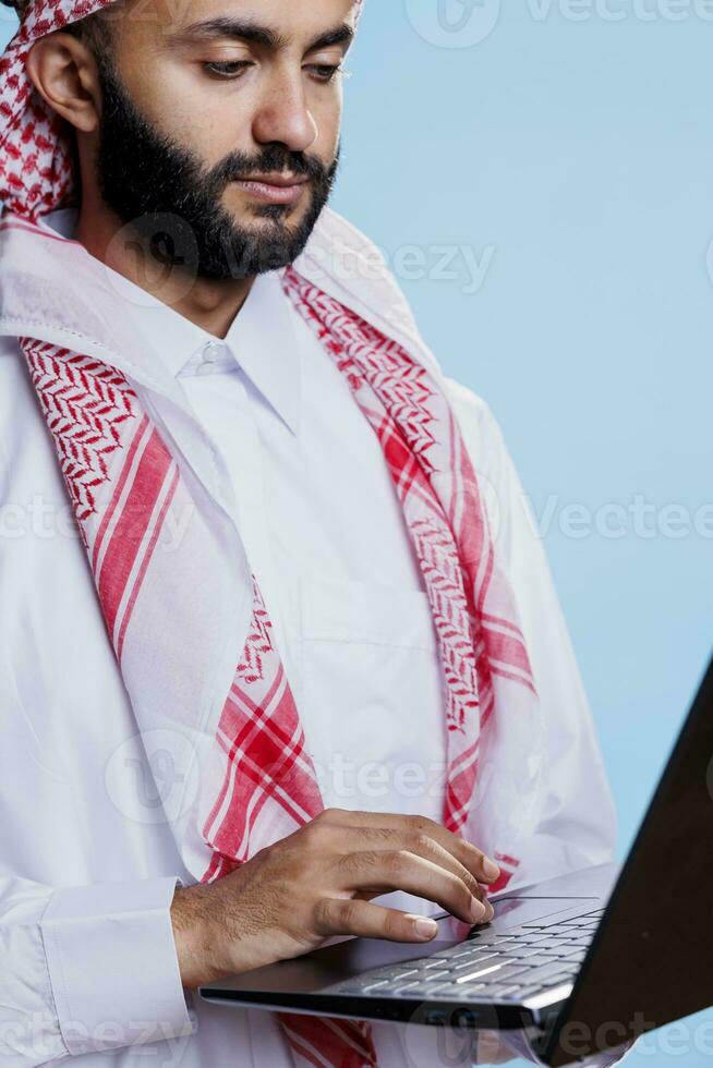 uomo vestito nel tradizionale musulmano thobe e utilizzando il computer portatile touchpad avvicinamento. arabo persona indossare islamico abbigliamento, Tenere portatile computer e scorrimento pagina mentre navigazione Internet foto