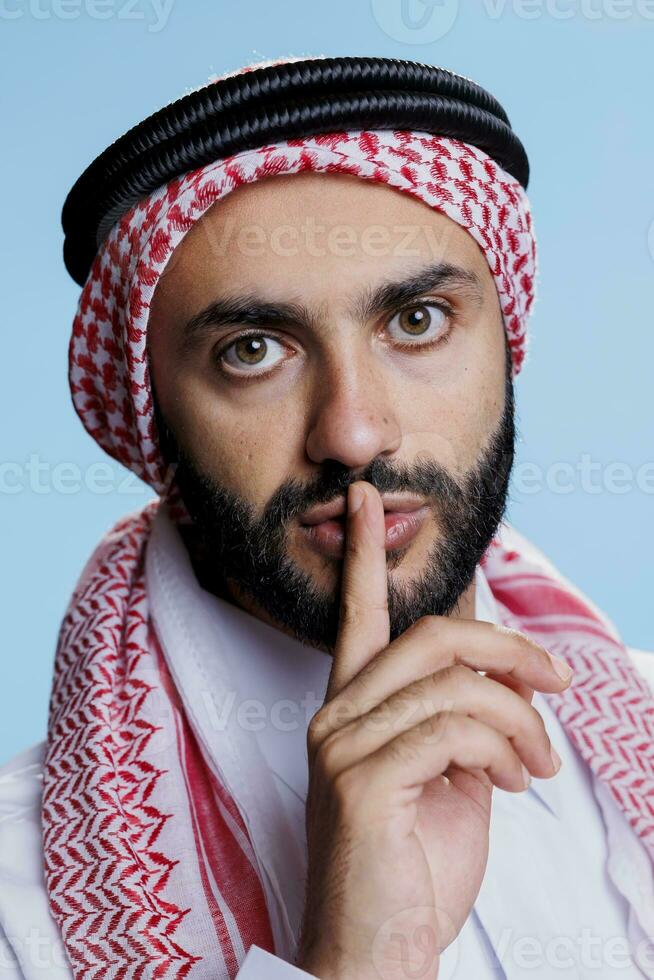 uomo indossare musulmano foulard segnalazione per silenzio con zittito dito per labbra avvicinamento ritratto. arabo nel copricapo con corda gruppo musicale mostrando silenzio gesto e guardare a telecamera con fiducioso espressione foto