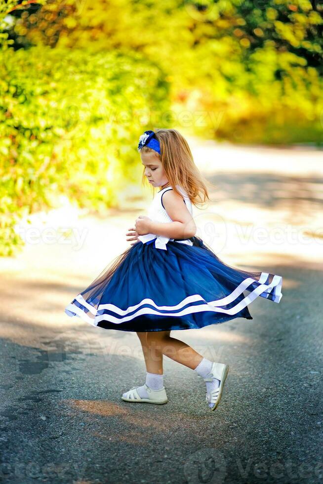 affascinante piccolo ragazza nel un' blu lungo vestito è danza e vortici nel un' soleggiato parco. bambino Filatura nel un' blu gonna. il poco ragazza passeggiate nel il cortile su un' sfondo di verde cespugli illuminato di il sole. foto