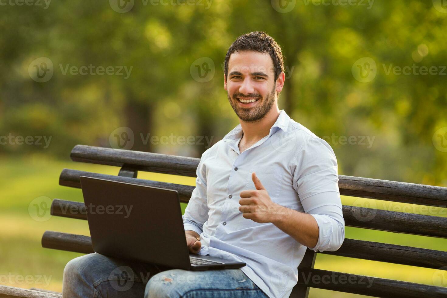 giovane uomo d'affari è seduta a il parco e utilizzando il computer portatile e mostrando pollice su foto