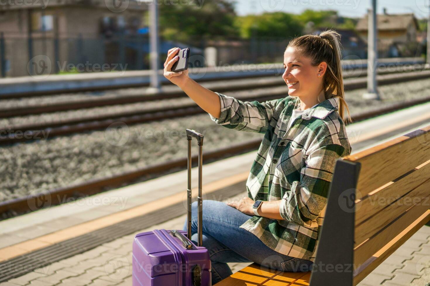 contento donna mostrando pollice su mentre assunzione autoscatto con mobile Telefono su un' treno stazione foto