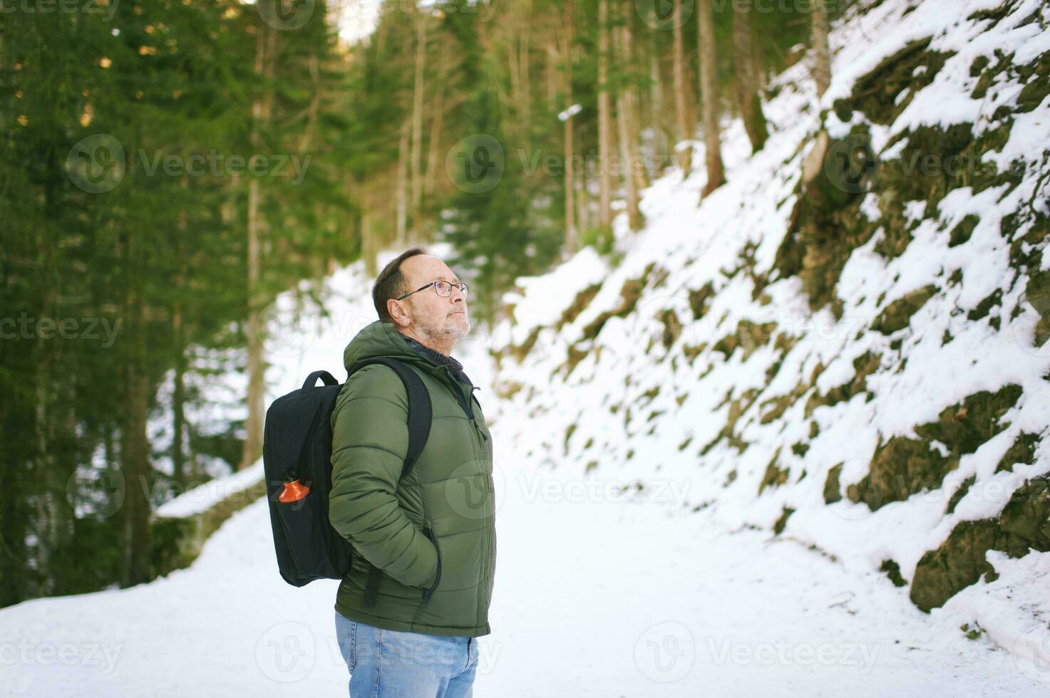 all'aperto ritratto di mezzo età 55 - 60 anno vecchio uomo escursioni a piedi nel inverno foresta, indossare caldo giacca e nero zaino foto