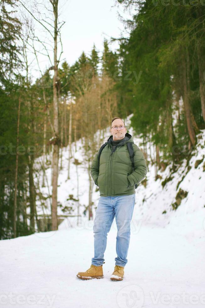 all'aperto ritratto di mezzo età 55 - 60 anno vecchio uomo escursioni a piedi nel inverno foresta, indossare caldo giacca e nero zaino foto