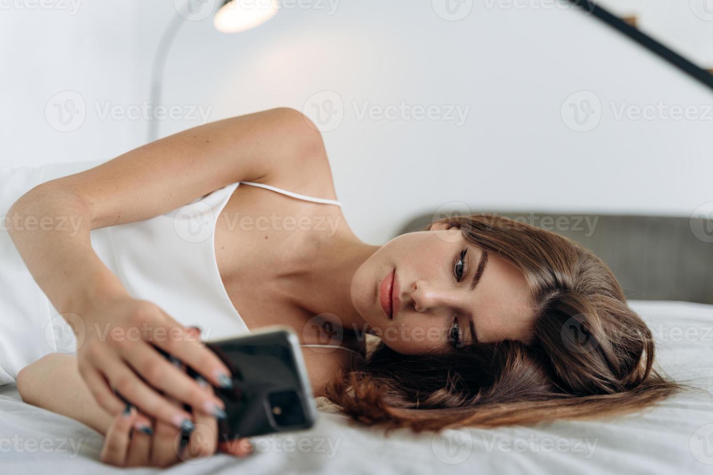 la giovane donna consapevole è sdraiata a letto dopo essersi svegliata e aver usato internet sullo smartphone. ragazza romantica e calma che guarda lo schermo del suo dispositivo foto