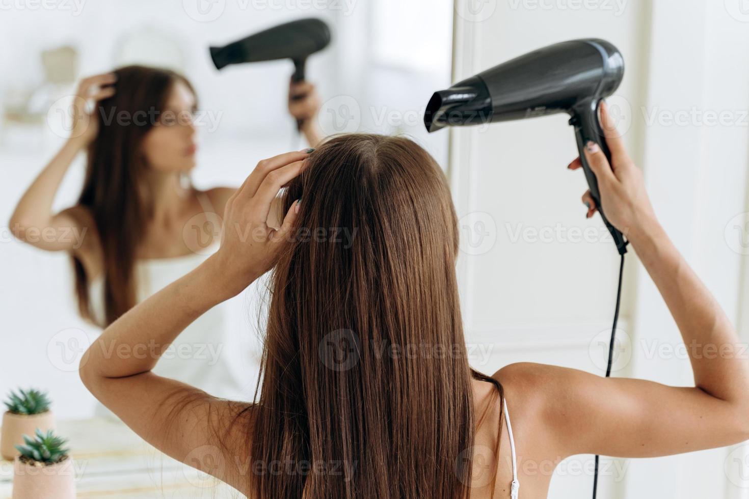 bella giovane donna con un asciugacapelli seduto davanti allo specchio e guardando il suo riflesso mentre si asciuga i capelli. cura per il concetto di chevelure foto