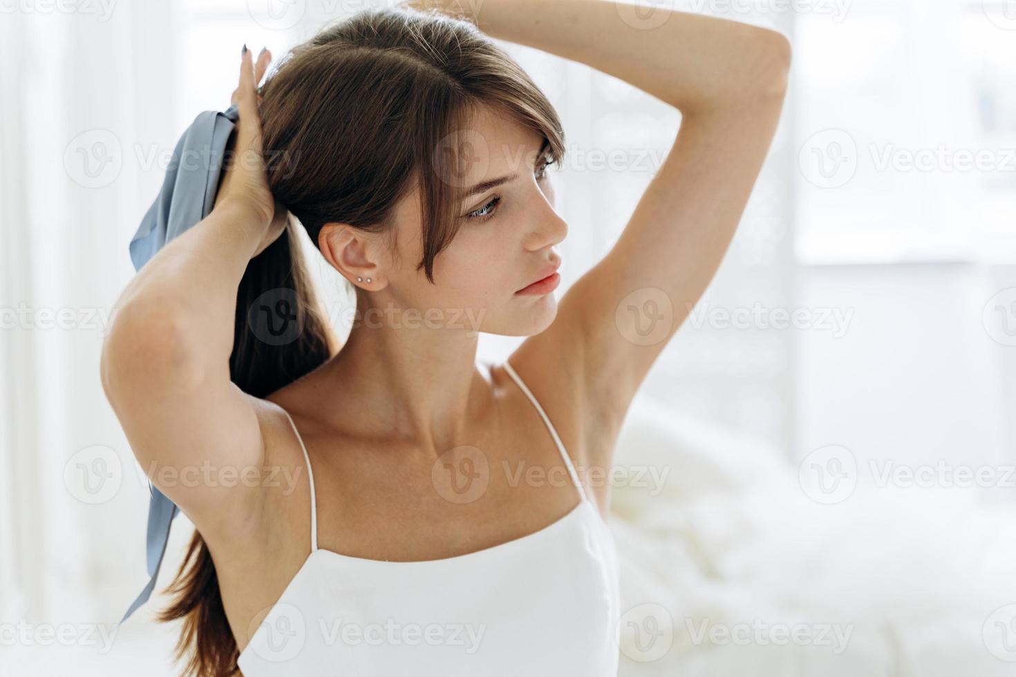 donna attraente che si spazzola i lunghi capelli castani in bagno al mattino tenendoli in una mano in una coda di cavallo mentre si prepara a legare un fiocco foto