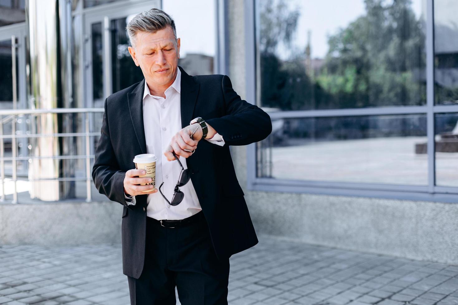 uomo d'affari senior che controlla il tempo sull'orologio in mano in città, con in mano occhiali da sole e bicchiere da caffè foto