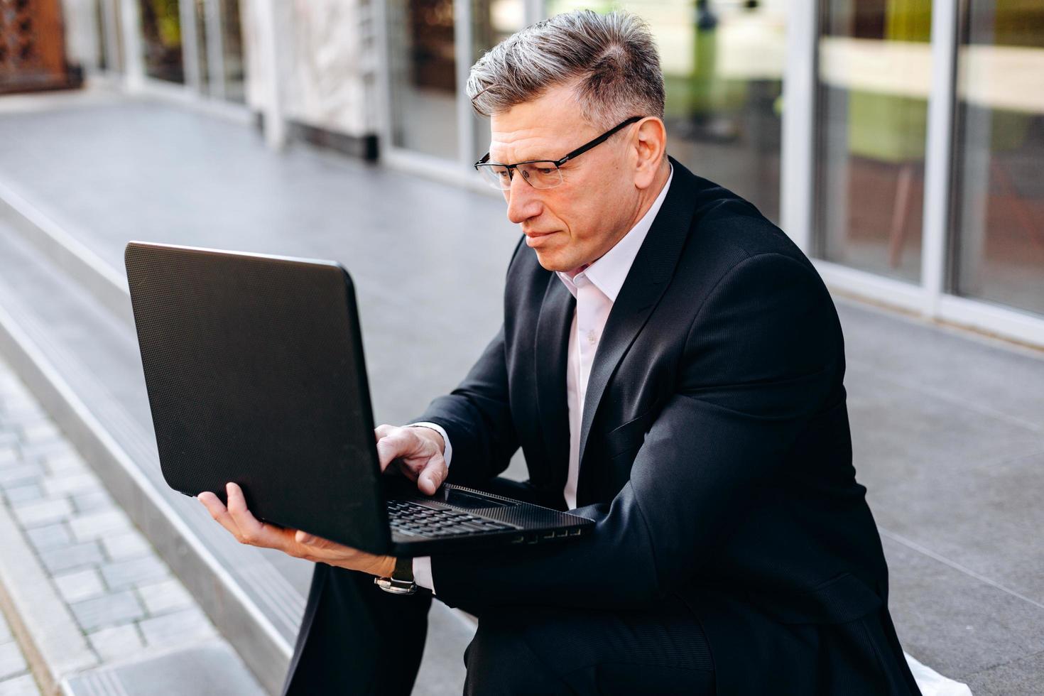 ritratto di uomo anziano in tuta seduto sul marciapiede e in possesso di un computer portatile e digitando all'aperto. - Immagine foto