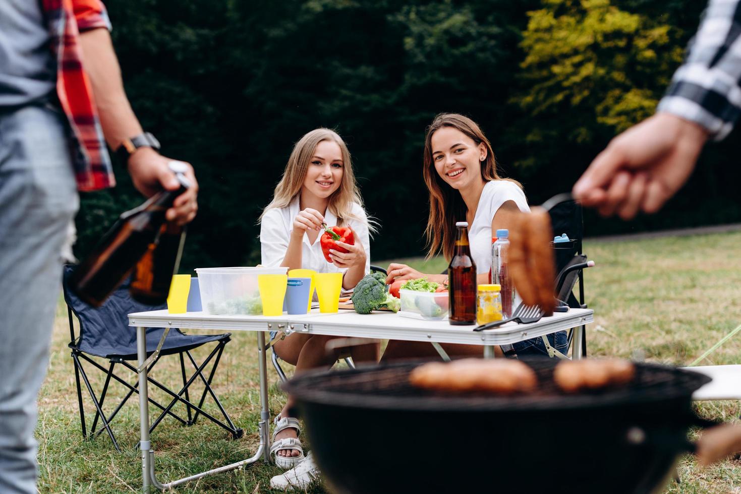 donne sorridenti sedute al tavolo con cibo fresco e guardando il barbecue foto