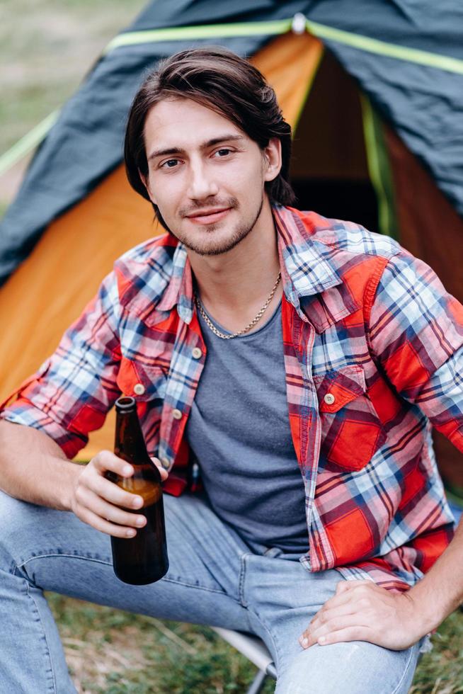 un ragazzo seduto accanto alla tenda con una bottiglia di birra e sorride guardando la telecamera foto