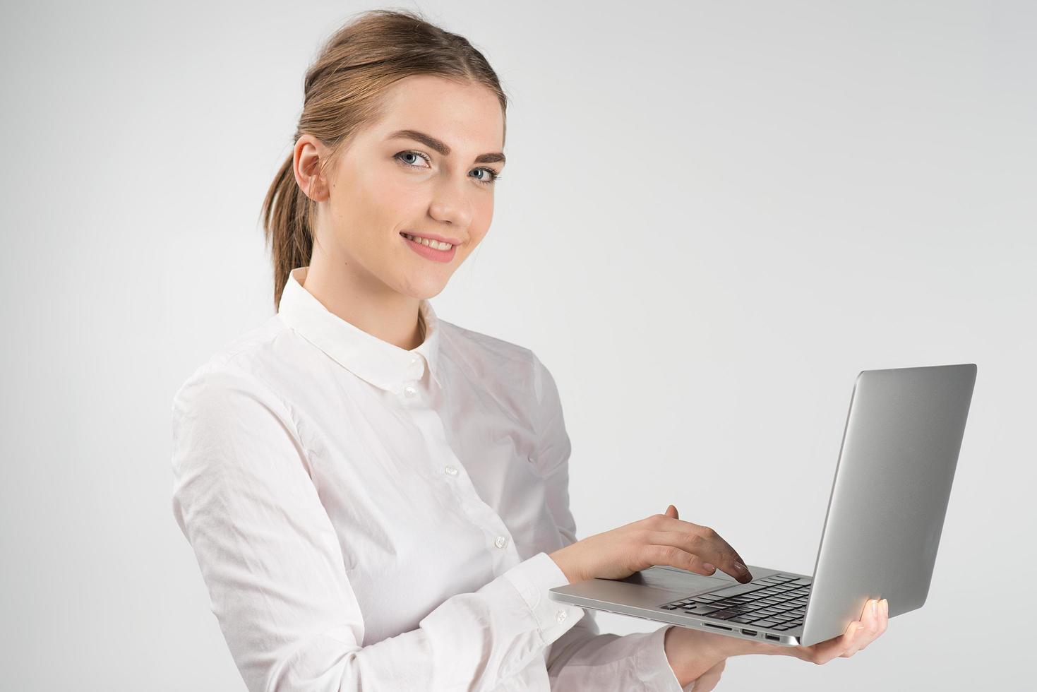 donna d'affari sorridente in camicia bianca che tiene in mano il computer portatile e mentre è in piedi e guarda la telecamera foto