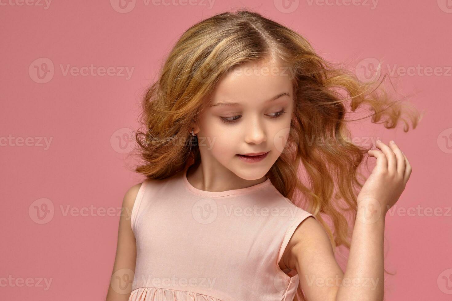 poco ragazza con un' biondo Riccio capelli, nel un' rosa vestito è in posa per il telecamera foto