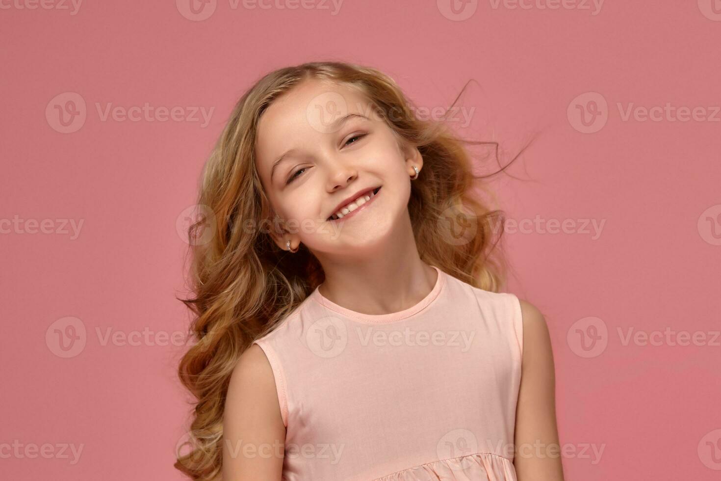 poco ragazza con un' biondo Riccio capelli, nel un' rosa vestito è in posa per il telecamera foto