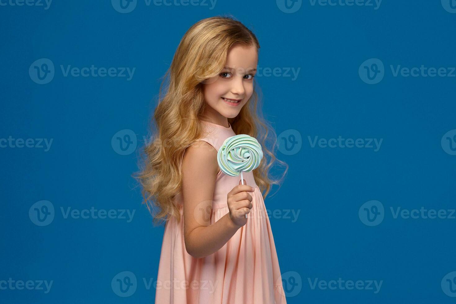 alla moda poco ragazza nel un' rosa vestire, con un' caramella nel sua mani, in piedi su un' blu sfondo. foto