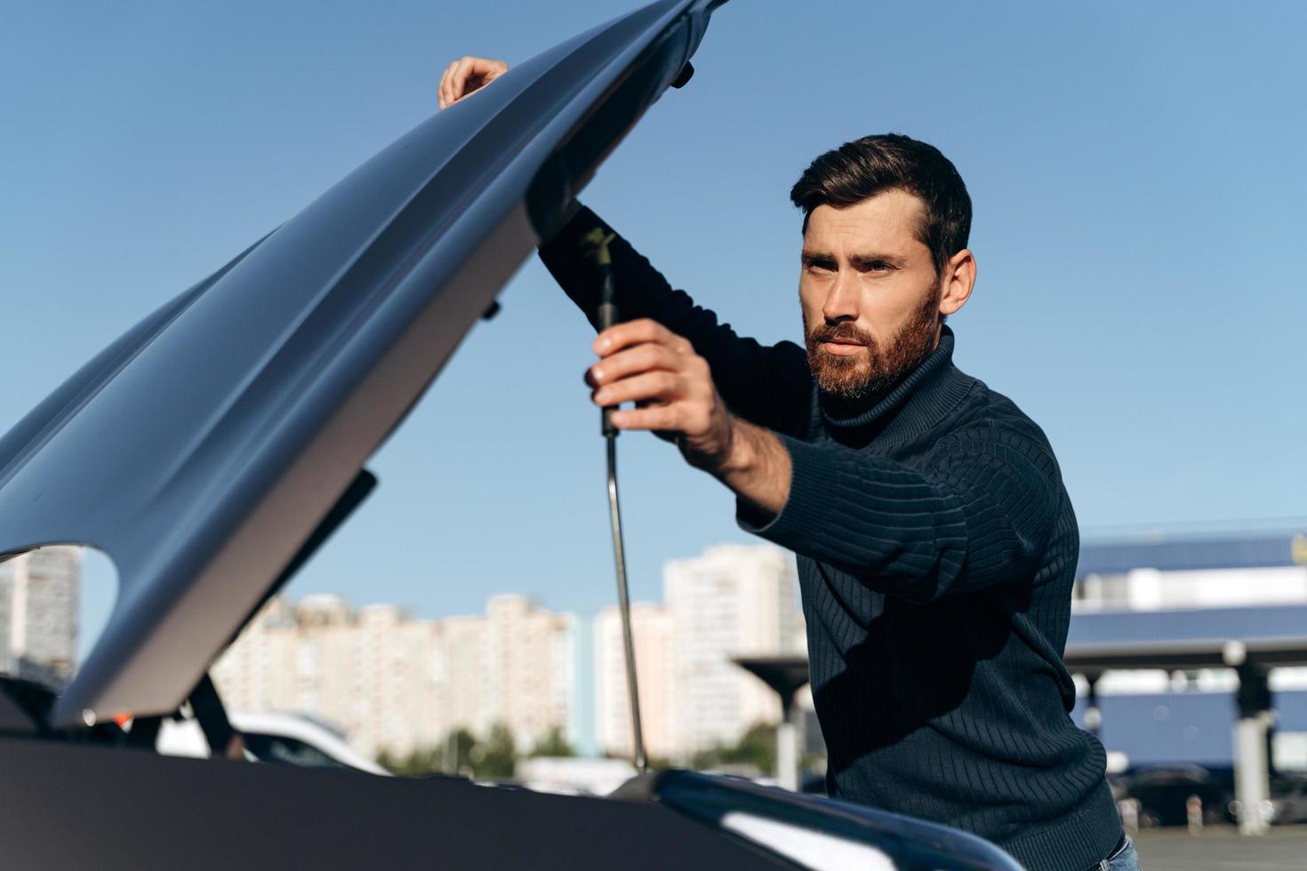 giovane che guarda sotto il cofano dell'auto in panne. guasto alla macchina. giovane concentrato cerca di riparare il motore, guardando dentro stando in piedi all'aperto foto