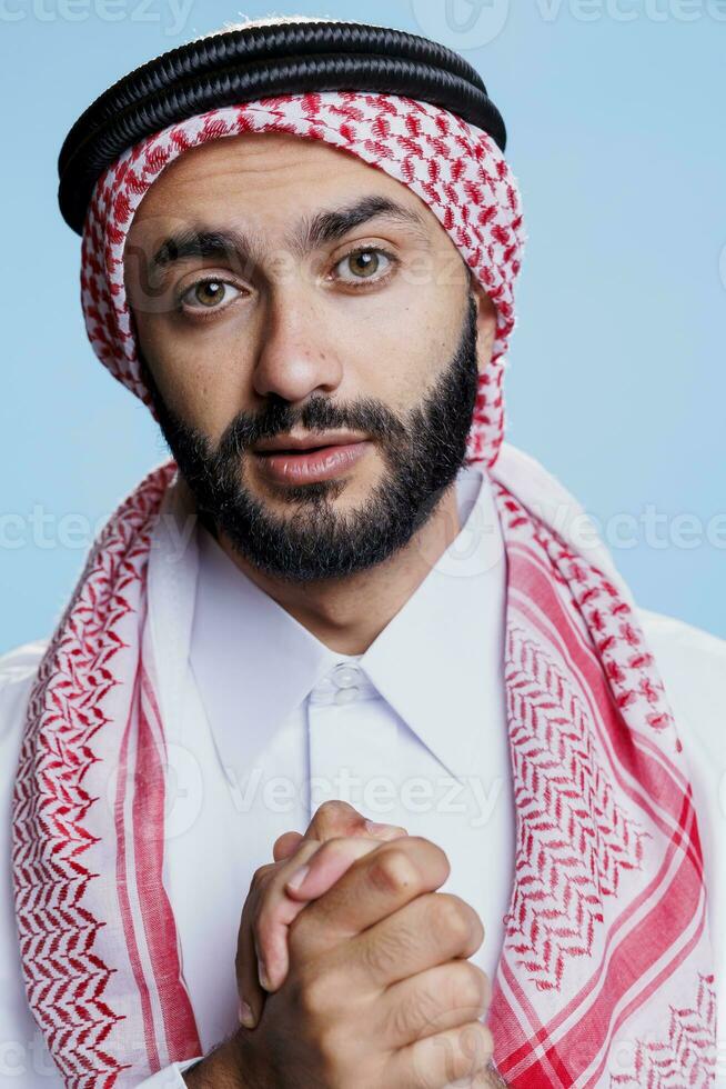 musulmano preghiera indossare tradizionale arabo Abiti mostrando religioso credenza e guardare a telecamera. islamico persona vestito nel ghutra foulard in posa nel preghiere posa nel studio foto