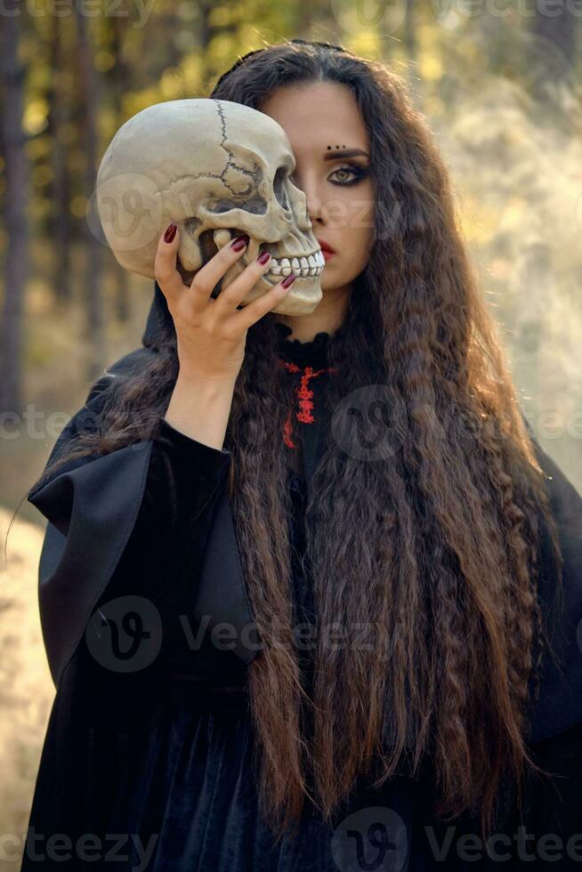 strega nel Nero, lungo vestito con capo e cappuccio. in posa nel Fumo pino foresta. Tenere un' cranio. incantesimi, Magia e stregoneria. avvicinamento. foto