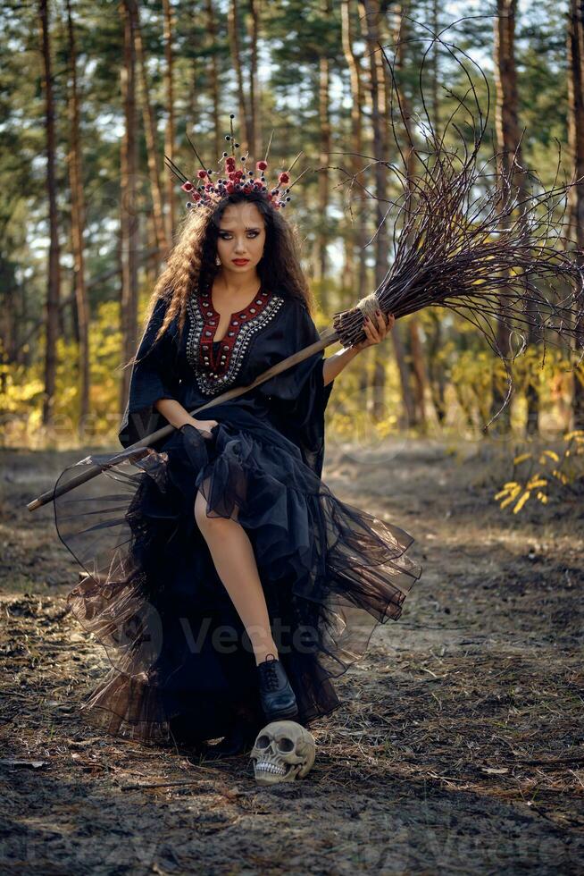 strega nel Nero, lungo vestire, con rosso corona nel sua lungo capelli. in posa con scopa e cranio nel pino foresta. incantesimi, Magia e stregoneria. pieno lunghezza. foto