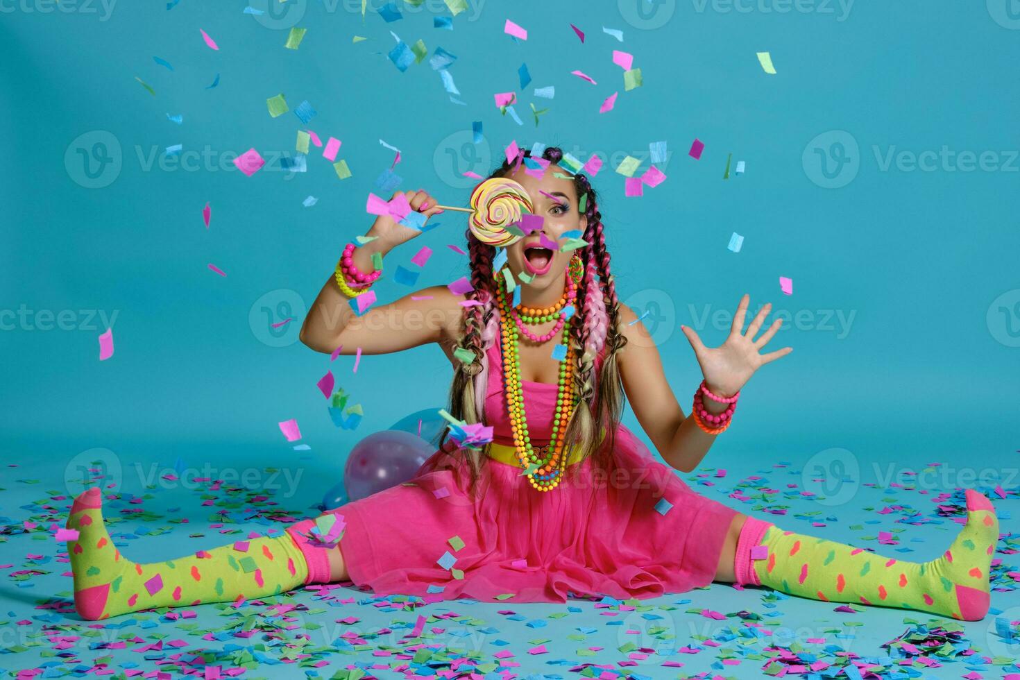 bello ragazza con un' multicolore trecce acconciatura e luminosa trucco, in posa nel studio con lecca-lecca, aria palloncini e coriandoli contro un' blu sfondo. foto