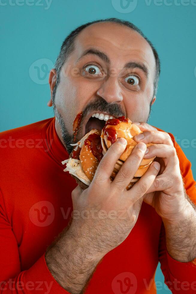 avvicinamento ritratto di un' di mezza età uomo con barba, vestito nel un' rosso dolcevita, in posa con hamburger contro un' blu sfondo. veloce cibo. foto