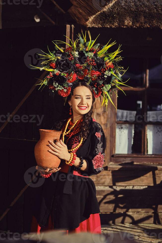 brunetta ragazza nel un' nero e rosso ucraino ricamato autentico nazionale costume e un' ghirlanda di fiori è in posa Tenere un' brocca e in piedi a il cancello. foto