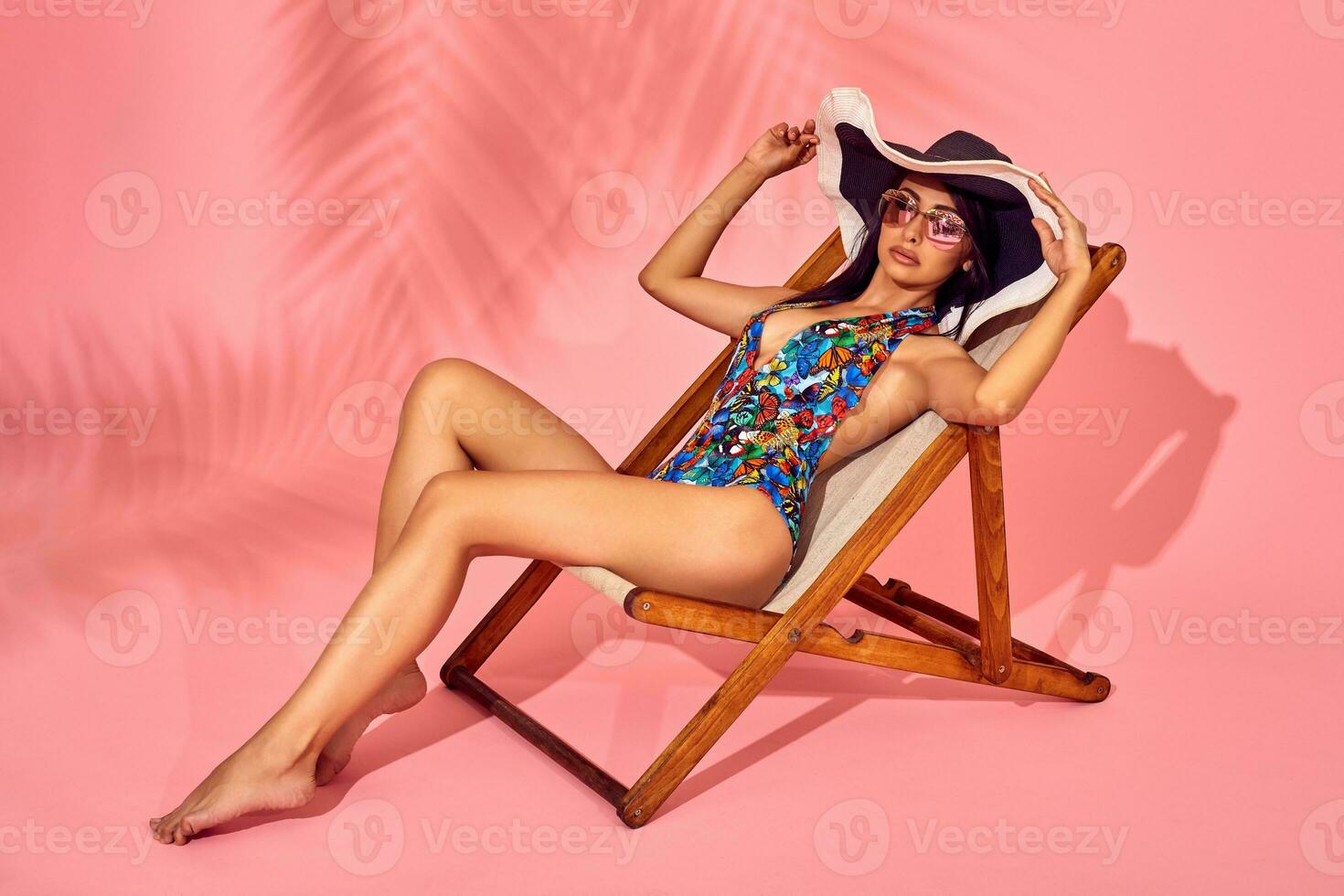 estate stile di vita moda ritratto di giovane sbalorditivo donna su un' sala sedia, rosa sfondo, studio sparo. indossare elegante occhiali da sole, bikini. foto