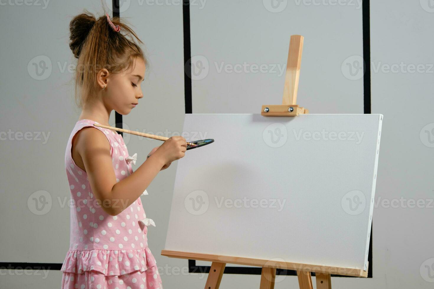 poco ragazza artista nel un' rosa vestito è in piedi dietro a cavalletto e pittura con spazzola su tela a arte studio con bianca muri. medio avvicinamento sparo. foto