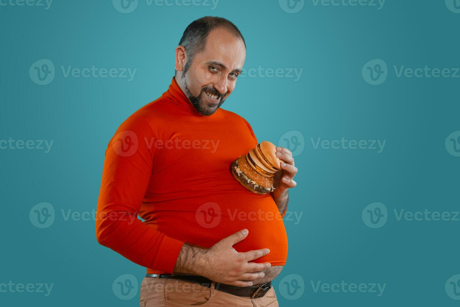 avvicinamento ritratto di un' di mezza età uomo con barba, vestito nel un' rosso dolcevita, in posa con hamburger contro un' blu sfondo. veloce cibo. foto