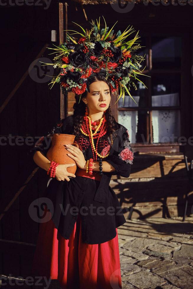 brunetta ragazza nel un' nero e rosso ucraino ricamato autentico nazionale costume e un' ghirlanda di fiori è in posa in piedi a il cancello. foto