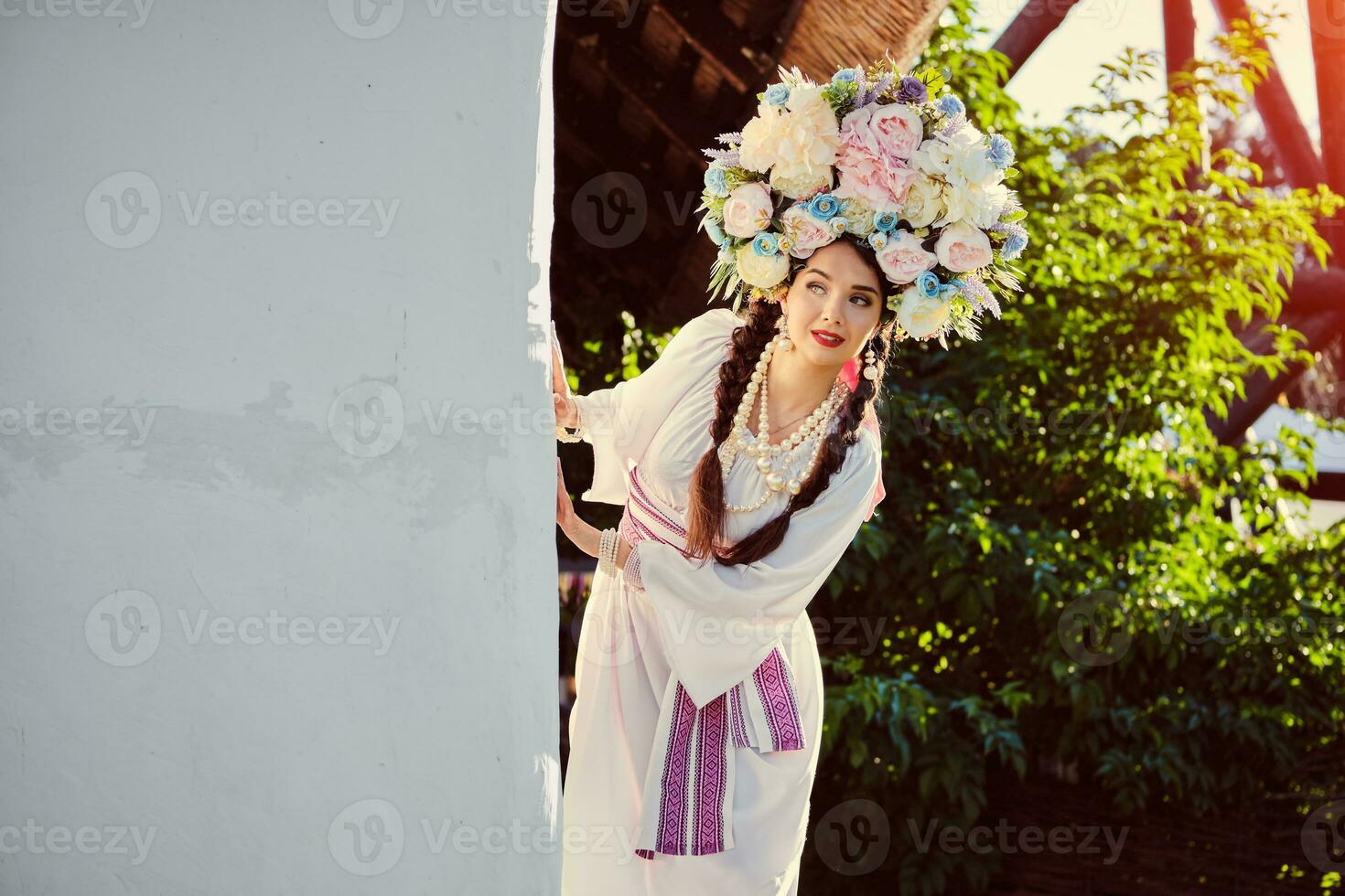 brunetta ragazza nel un' bianca ucraino autentico nazionale costume e un' ghirlanda di fiori è in posa contro un' bianca capanna. foto