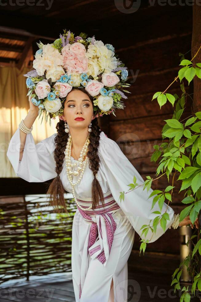 brunetta ragazza nel un' bianca ucraino autentico nazionale costume e un' ghirlanda di fiori è in posa contro un' verde cortile. avvicinamento. foto