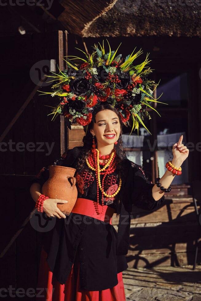 brunetta ragazza nel un' nero e rosso ucraino ricamato autentico nazionale costume e un' ghirlanda di fiori è in posa in piedi a il cancello. foto