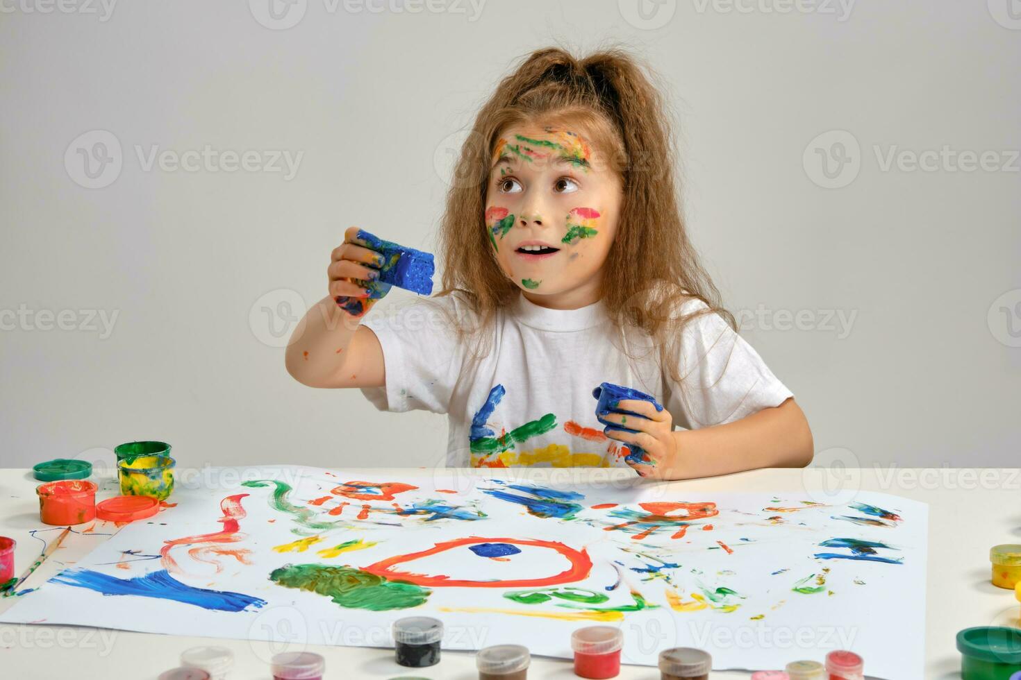 poco ragazza nel bianca maglietta seduta a tavolo con che uomo e vernici, disegno su esso, in posa con dipinto viso, mani. isolato su bianca. avvicinamento. foto