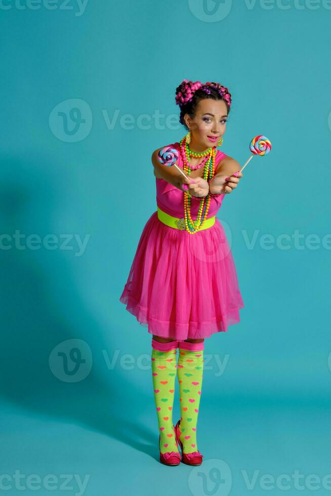 bello ragazza con un' multicolore trecce acconciatura e luminosa trucco, in posa nel studio contro un' blu sfondo, Tenere un' lecca-lecca nel sua mano. foto