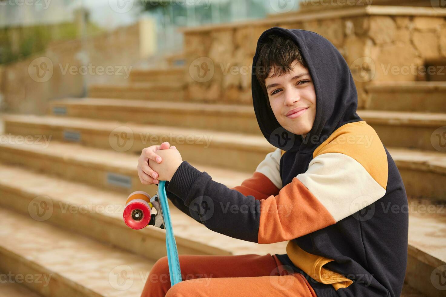 caucasico adolescente fricchettone ragazzo seduta su passaggi sfondo, vestito nel un' nero felpa, Tenere skateboard, guardare a telecamera foto