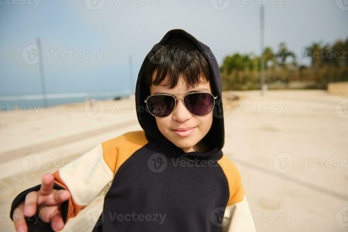 Multi etnico adolescente nel occhiali da sole e nero felpa con cappuccio con cappuccio su il suo testa, sorridente a telecamera equitazione skateboard all'aperto foto