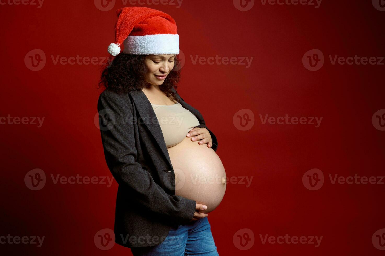 affascinante mezzo anziano incinta donna attenzione un' Santa cappello, Tenere sua nudo gonfiarsi, sorridente, isolato al di sopra di rosso sfondo foto