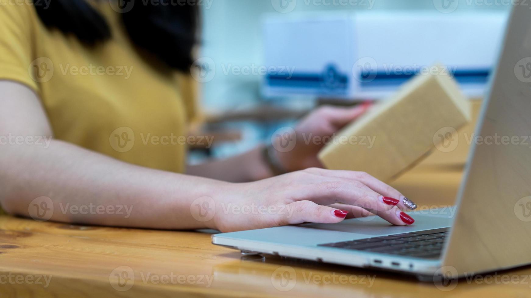una donna che vende online utilizza un laptop e tiene in mano una cassetta dei pacchi per controllare le informazioni sui clienti. foto