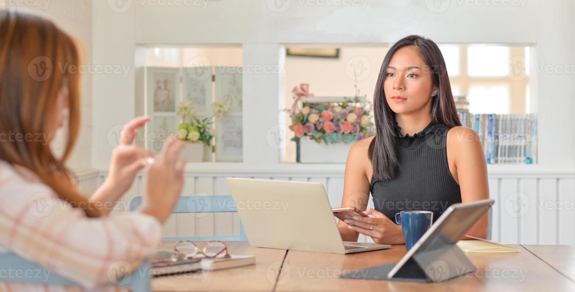 la giovane donna tiene uno smartphone in mano e un laptop con tablet sul tavolo. lei e la sua amica stanno preparando un progetto per presentare un cliente. foto