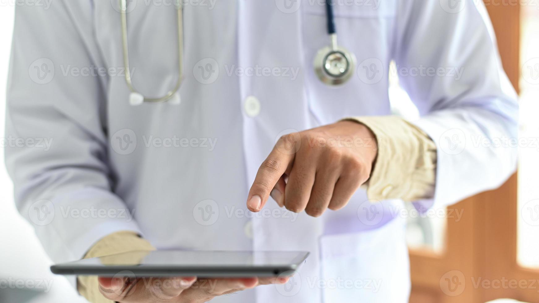 primo piano di un medico che punta a un tablet, un uomo in un camice medico con uno stetoscopio che fa funzionare il tablet. foto
