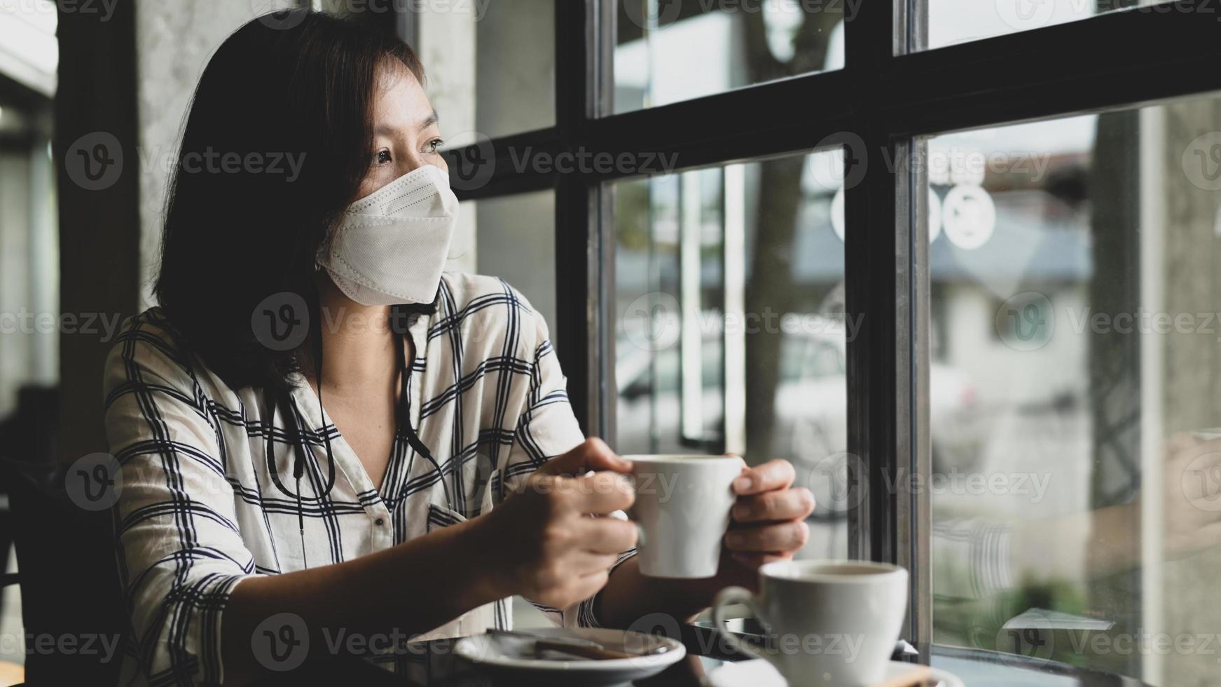 una donna che indossa una maschera medica siede in una caffetteria guardando fuori dalla finestra con occhi tristi. foto
