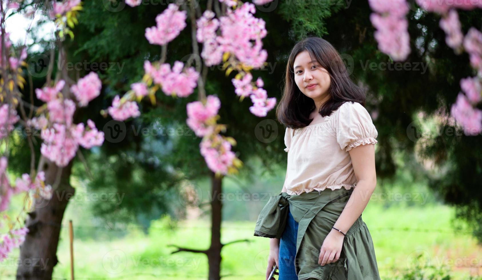 giovane ragazza asiatica guarda la telecamera e scatta foto sotto un albero di fiori di ciliegio.