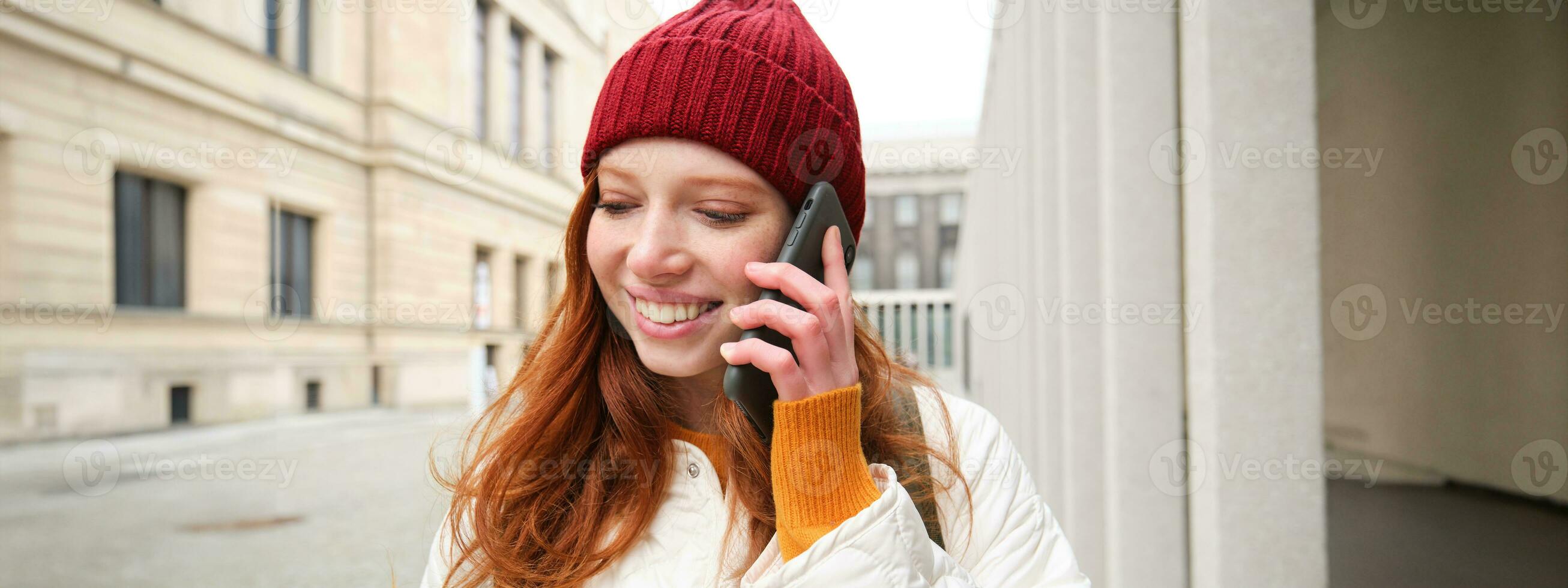 mobile banda larga e le persone. sorridente giovane testa Rossa donna passeggiate nel cittadina e parla su mobile Telefono, chiamata amico su smartphone, utilizzando Internet per rendere un' chiamata all'estero foto