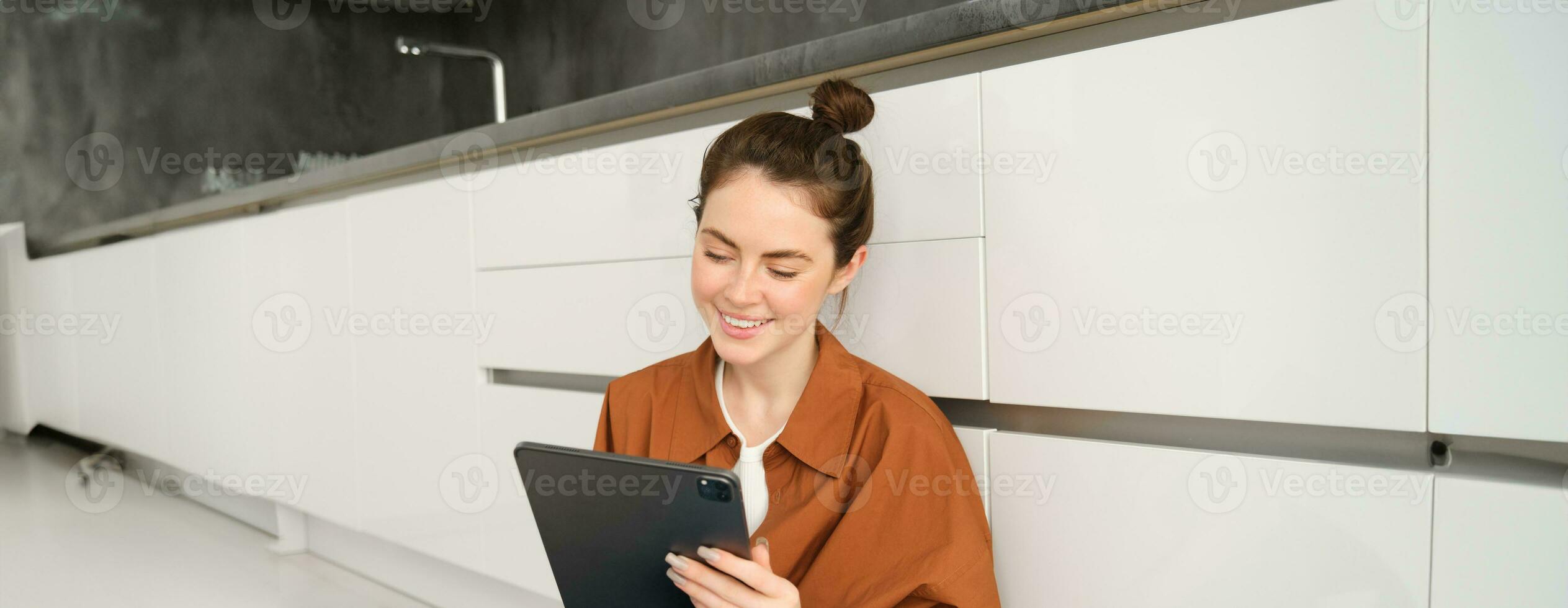 ritratto di giovane bellissimo donna seduta su cucina pavimento con digitale tavoletta, navigazione notizia alimentazione, sociale media App su gadget, sorridente e guardare contento foto