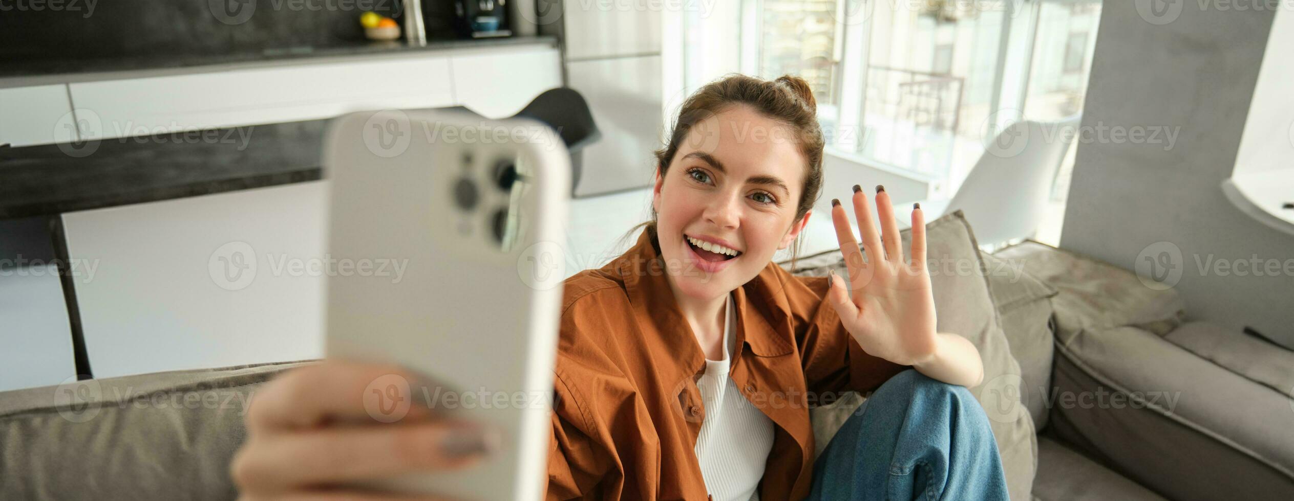 amichevole sorridente giovane donna, seduta su divano e agitando mano a smartphone telecamera, video chat, parlando su mobile Telefono, si connette per in linea conversazione su applicazione foto