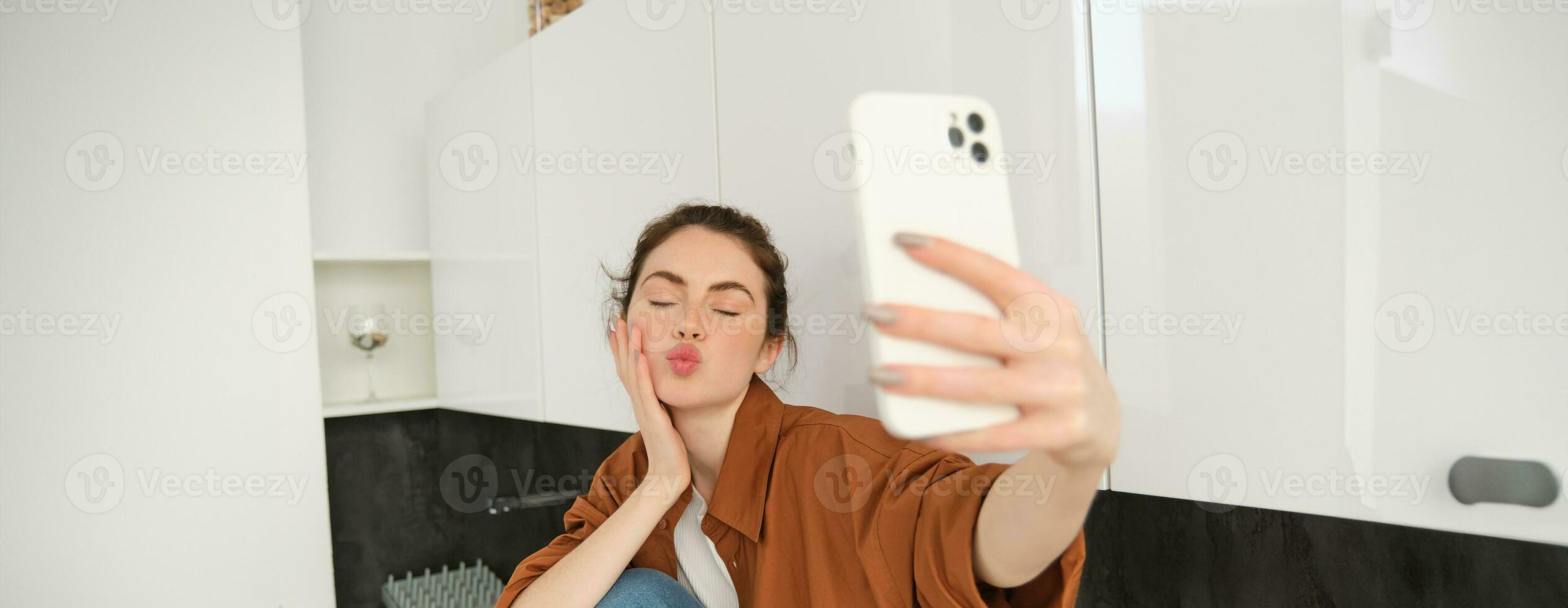 giovane carismatico ragazza prende selfie nel il cucina, in posa per foto, detiene mobile Telefono con esteso mano foto