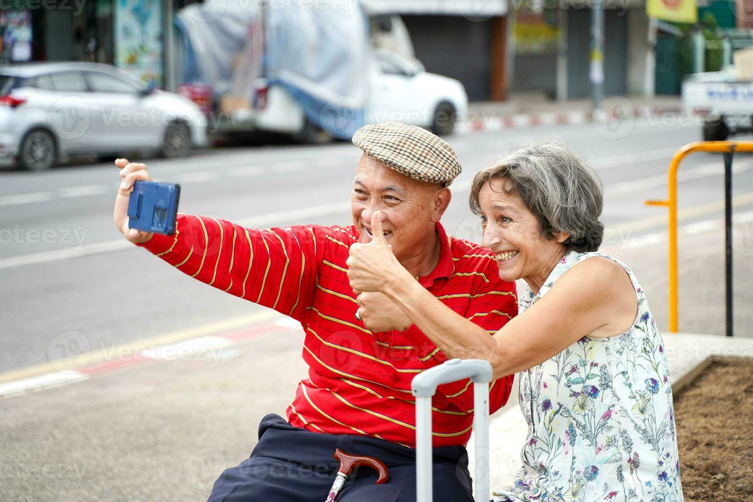 anziano Cinese turista con il suo amico europeo pose contento pollici su e prendere un' foto autoscatto su sfocato di città sfondo.